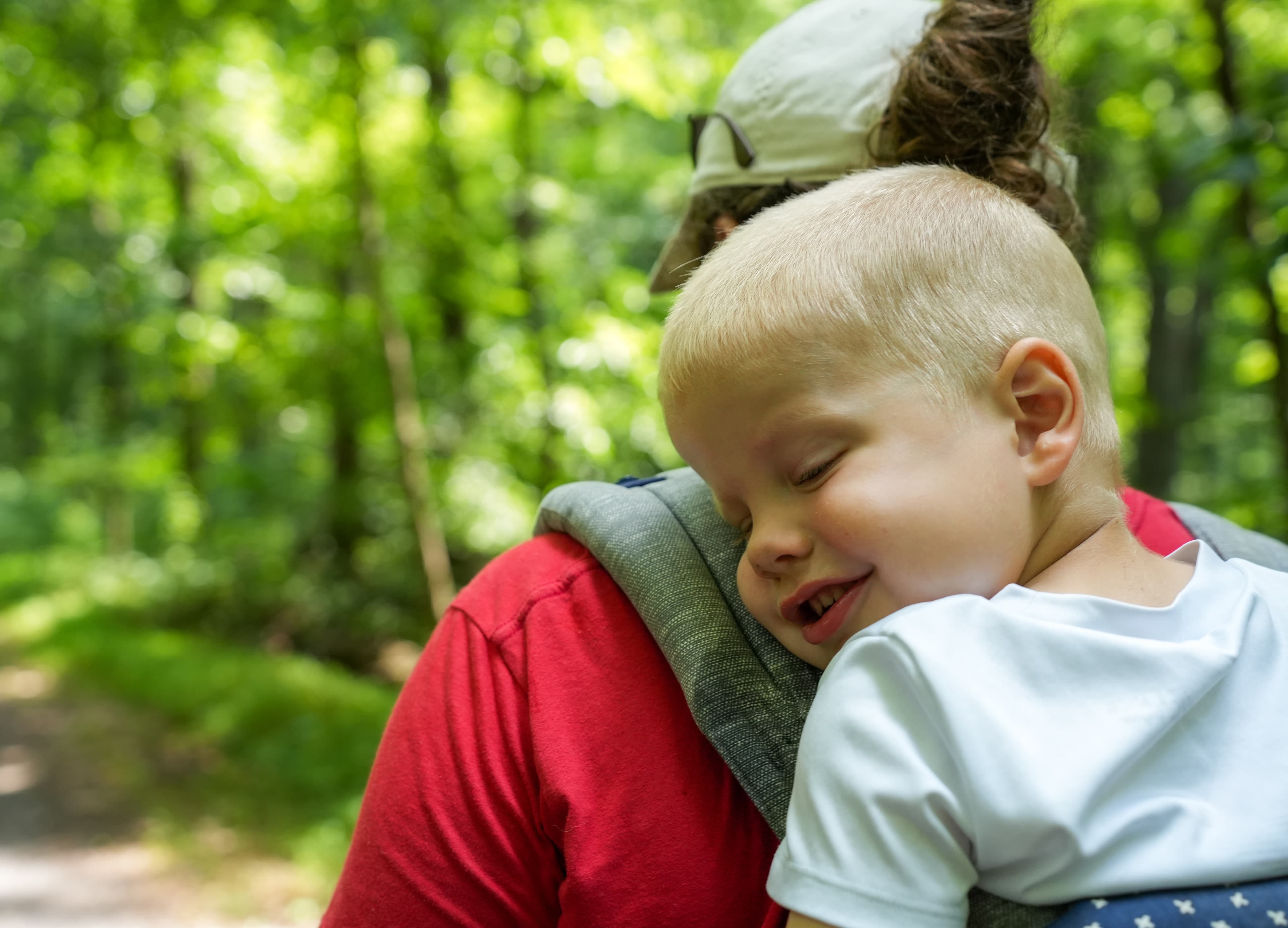 A toddler asleep on his mother's shoulder on a forest trail