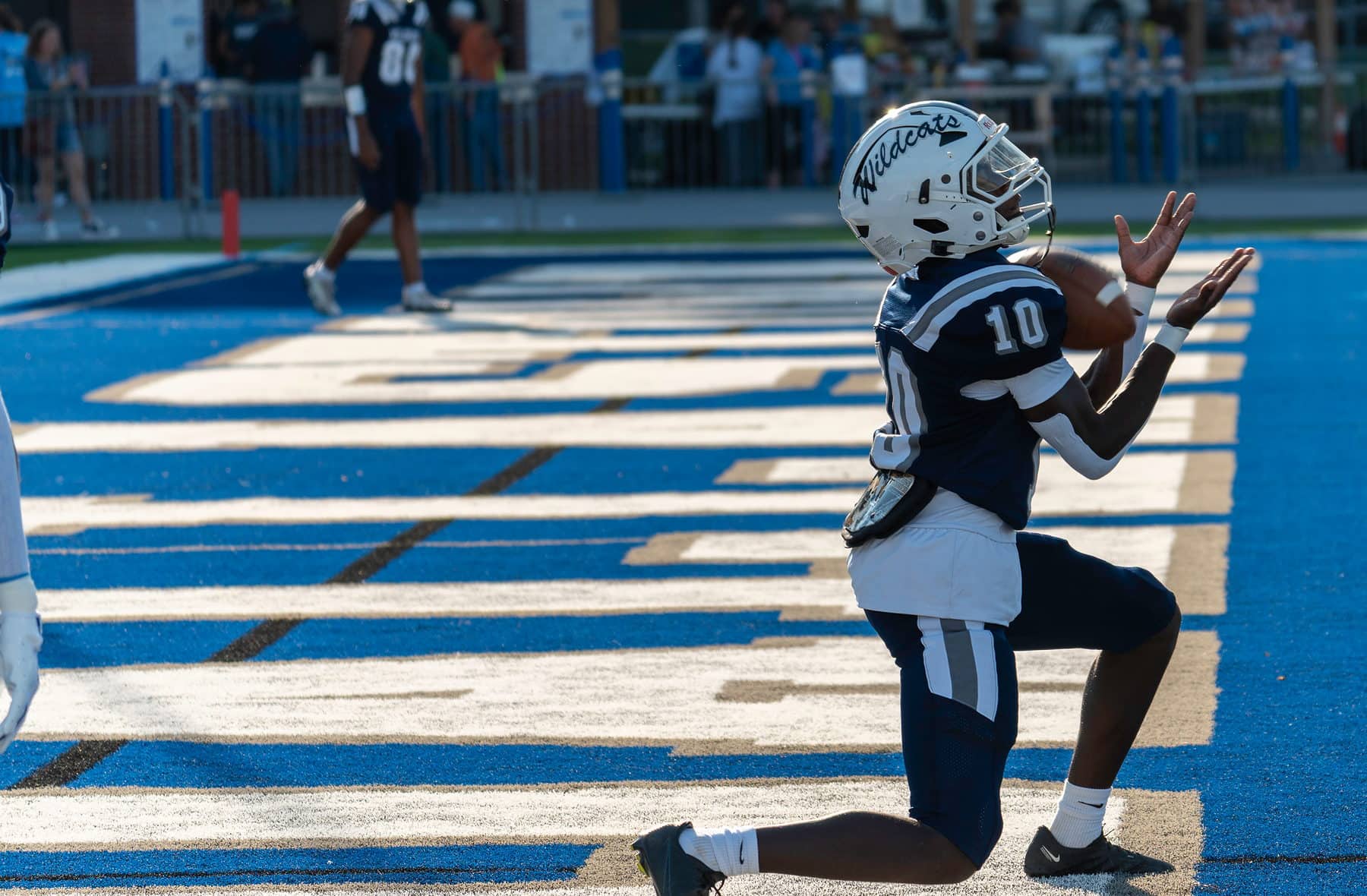 A high school receiver kneeling in the end zone after a touchdown, backlit by stadium lights